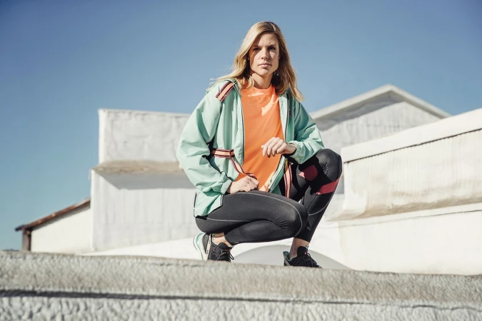 Female athlete in mint green jacket and black leggings squats on a concrete surface, set against a clear blue sky and minimalist white buildings.