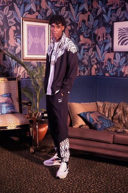 Full-length shot of a young adult man in a patterned tracksuit and sneakers in a styled interior with patterned wallpaper and furniture.
