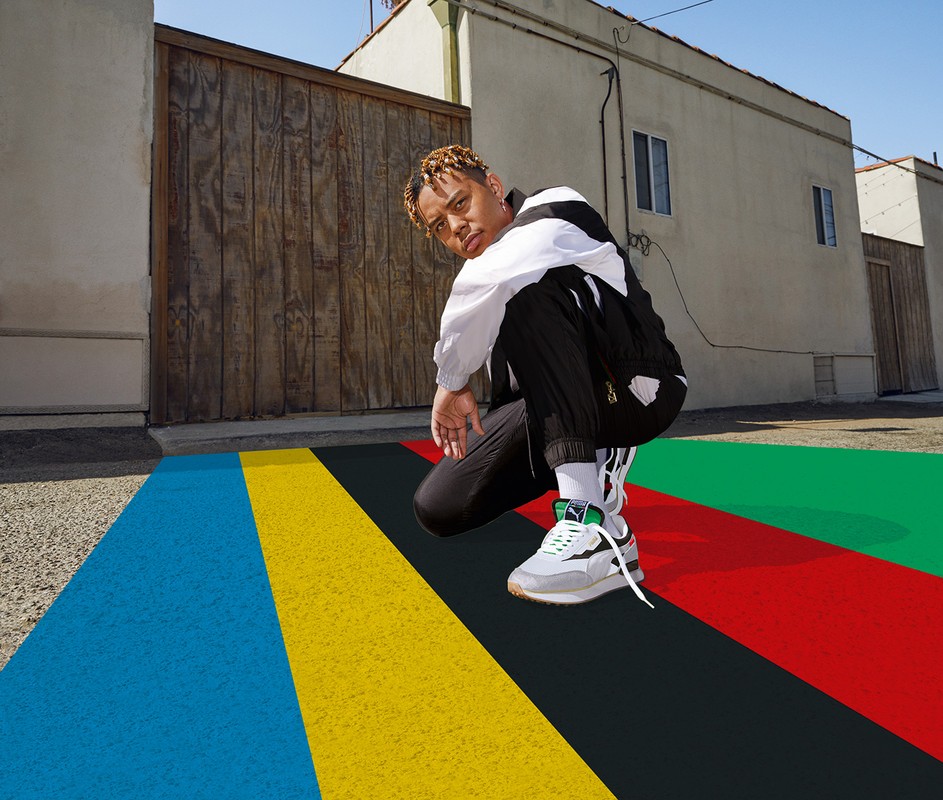 Young adult in black and white jacket and pants crouching on a brightly colored surface with sneakers and buildings in the background.