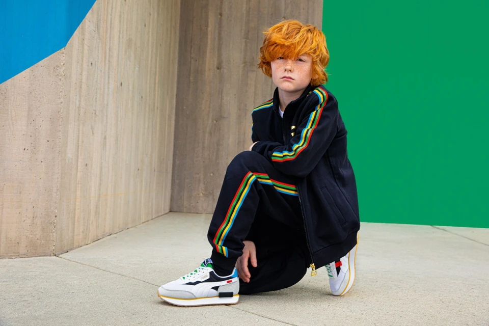 Boy with red hair and tracksuit squatting and looking at the camera in front of a blue, green, and concrete background.