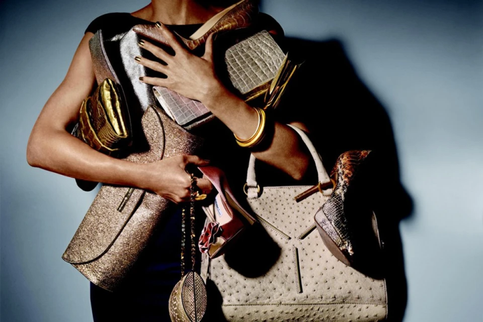Woman holding multiple handbags and high-heeled shoes in an indoor setting with blue gradient background, highlighted by studio lighting.