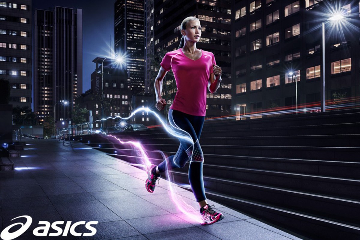 Woman runs at night on city sidewalk wearing pink athletic shirt and black leggings with digital light effects and Asics logo.