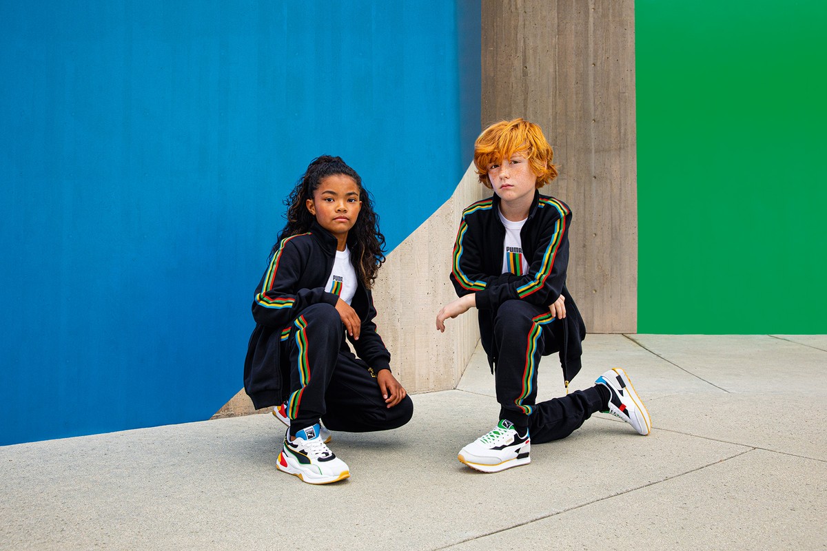 Two kids in matching black tracksuits and sneakers pose against a backdrop of bright blue, green, and grey concrete wall.