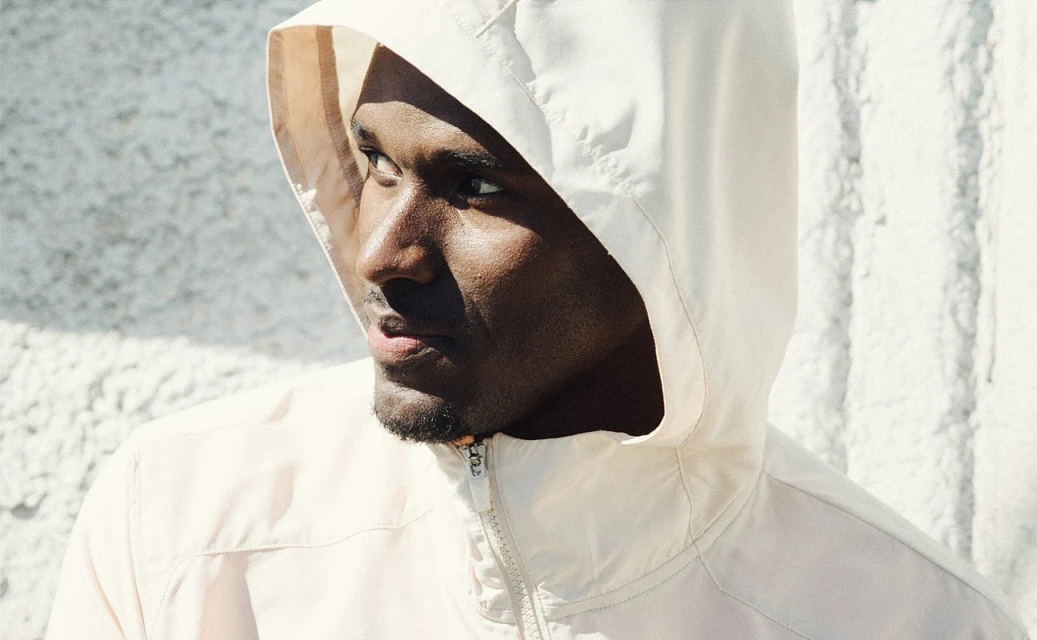 Close up of a man in a hooded jacket with a serious expression standing in front of a textured white wall bathed in bright sunlight.