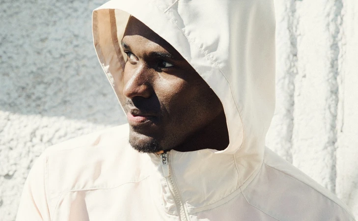 Close up of a man in a hooded jacket with a serious expression standing in front of a textured white wall bathed in bright sunlight.