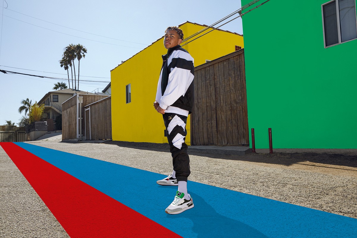 Man in Puma tracksuit stands outdoors against colorful buildings on a street painted with red, blue, and gray.