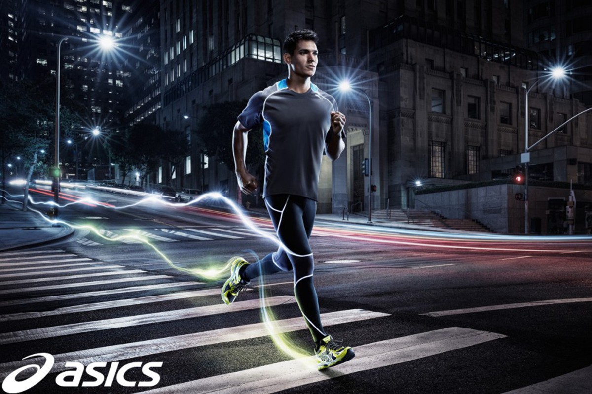 A man is running at night on a crosswalk in athletic wear with buildings and streetlights in the background, motion blur is apparent.
