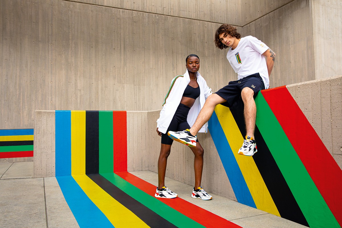 Two models showcase athletic wear in an urban setting, posing against a colorfully painted wall. One model sits, the other stands wearing Puma sneakers.