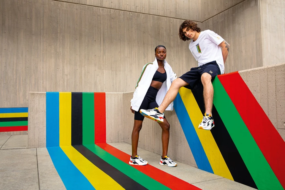 Two models showcase athletic wear in an urban setting, posing against a colorfully painted wall. One model sits, the other stands wearing Puma sneakers.