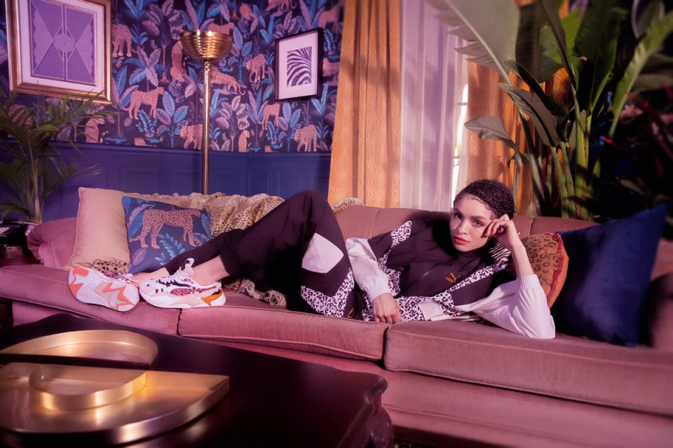 Woman in Puma tracksuit lounges on mauve couch in a living room with animal print wallpaper, decorative throw pillows, and athletic shoes.