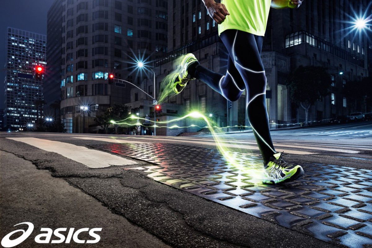 A man is running across a crosswalk in a city at night. He is wearing a neon green shirt, black leggings, and running shoes.