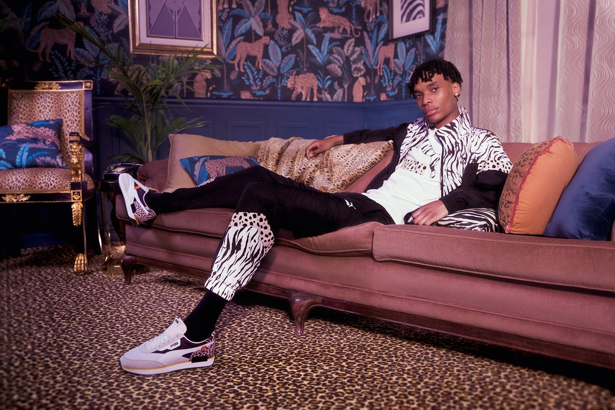 Adult male in athletic apparel with mixed zebra and leopard prints lounges on a couch in a room with patterned wallpaper and carpet.