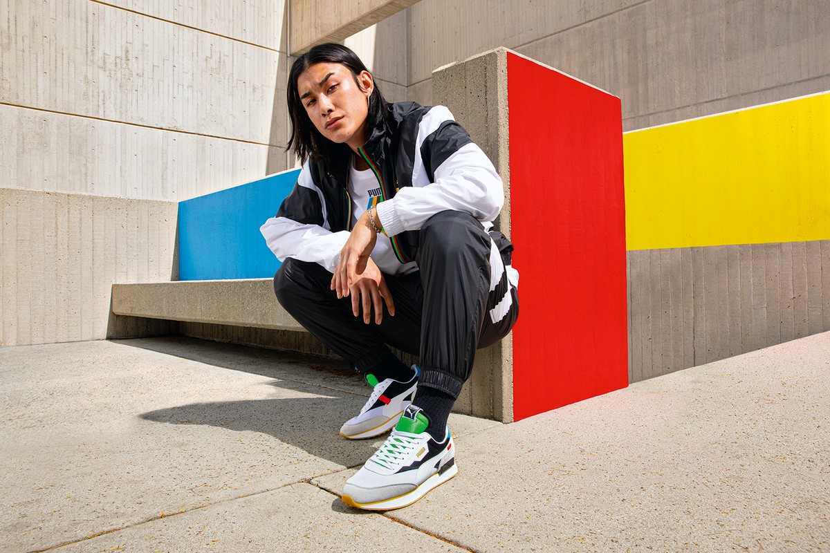 Man in athletic jacket and sweatpants crouching outdoors next to concrete and colorful painted surfaces, wearing Puma sneakers.
