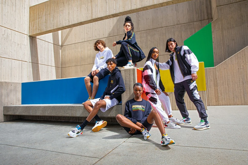Group of diverse adult fashion models in streetwear posing in an urban setting with concrete walls and colorful accents on a sunny day.
