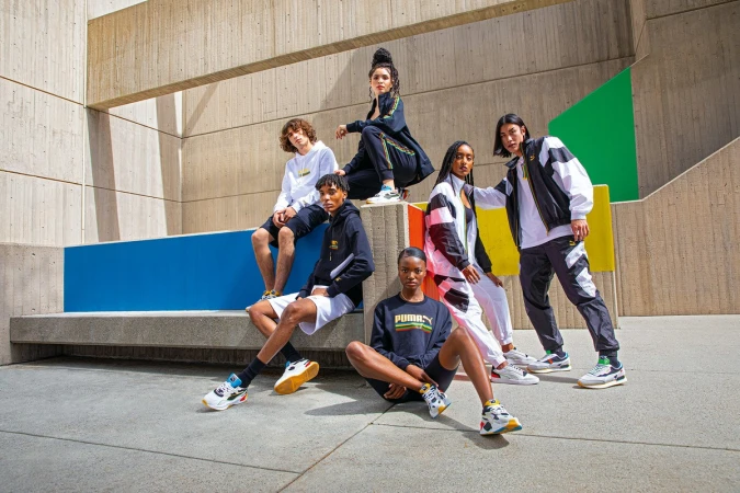 Group of diverse adult fashion models in streetwear posing in an urban setting with concrete walls and colorful accents on a sunny day.