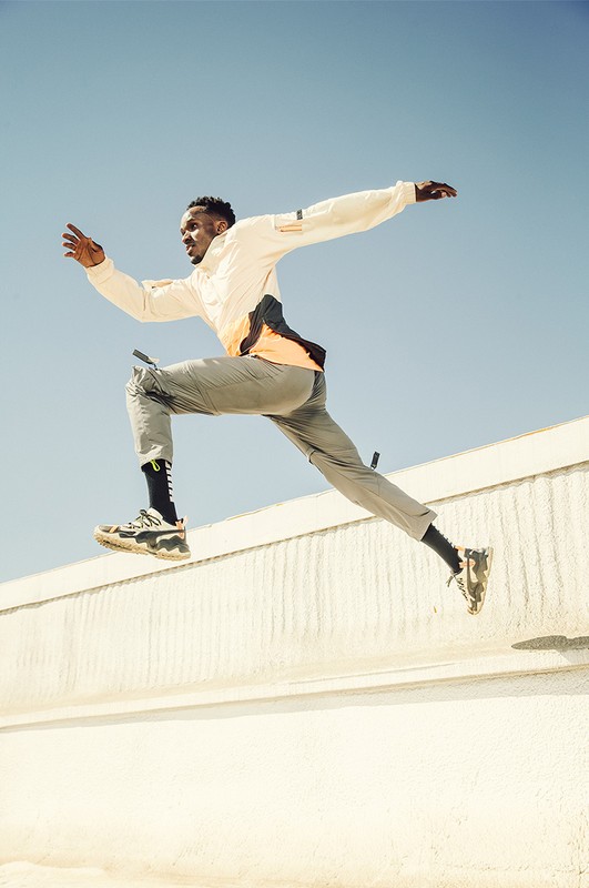 Athletic African American man leaps over a low wall on a bright sunny day, wearing sports clothing and sneakers, set against a clear blue sky.