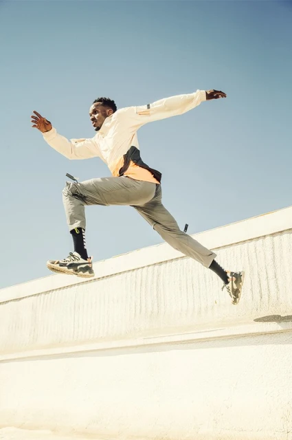 Athletic African American man leaps over a low wall on a bright sunny day, wearing sports clothing and sneakers, set against a clear blue sky.