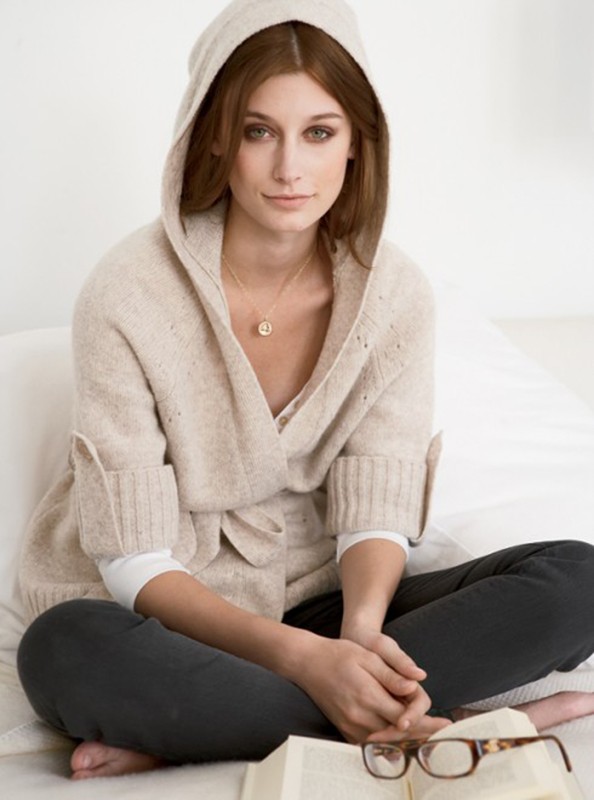 Adult female in a knitted hooded sweater sits cross-legged on a bed, with an open book and glasses in front of her, looking at the camera.