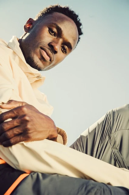 Low angle portrait of a dark-skinned male in athletic wear against a pale blue sky, focusing on his face and casual pose. 