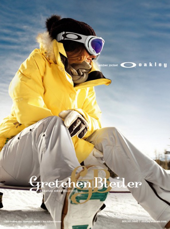 A woman in a yellow jacket, white ski pants, and Oakley goggles sits on snow against a blue sky, showcasing winter sports fashion.