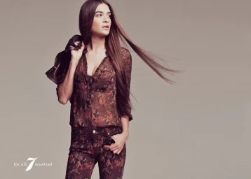 Model with long brown hair wearing a floral print blouse and matching trousers, holding a jacket over her shoulder against a grey background.