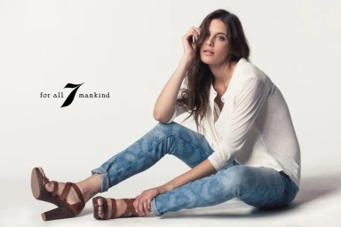 Woman seated on white studio floor wearing patterned blue jeans, white blouse, and brown wedge heels.