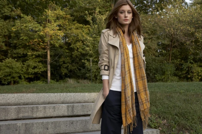 A woman wearing a beige trench coat and yellow plaid scarf standing outdoors near stone steps with trees in the background.