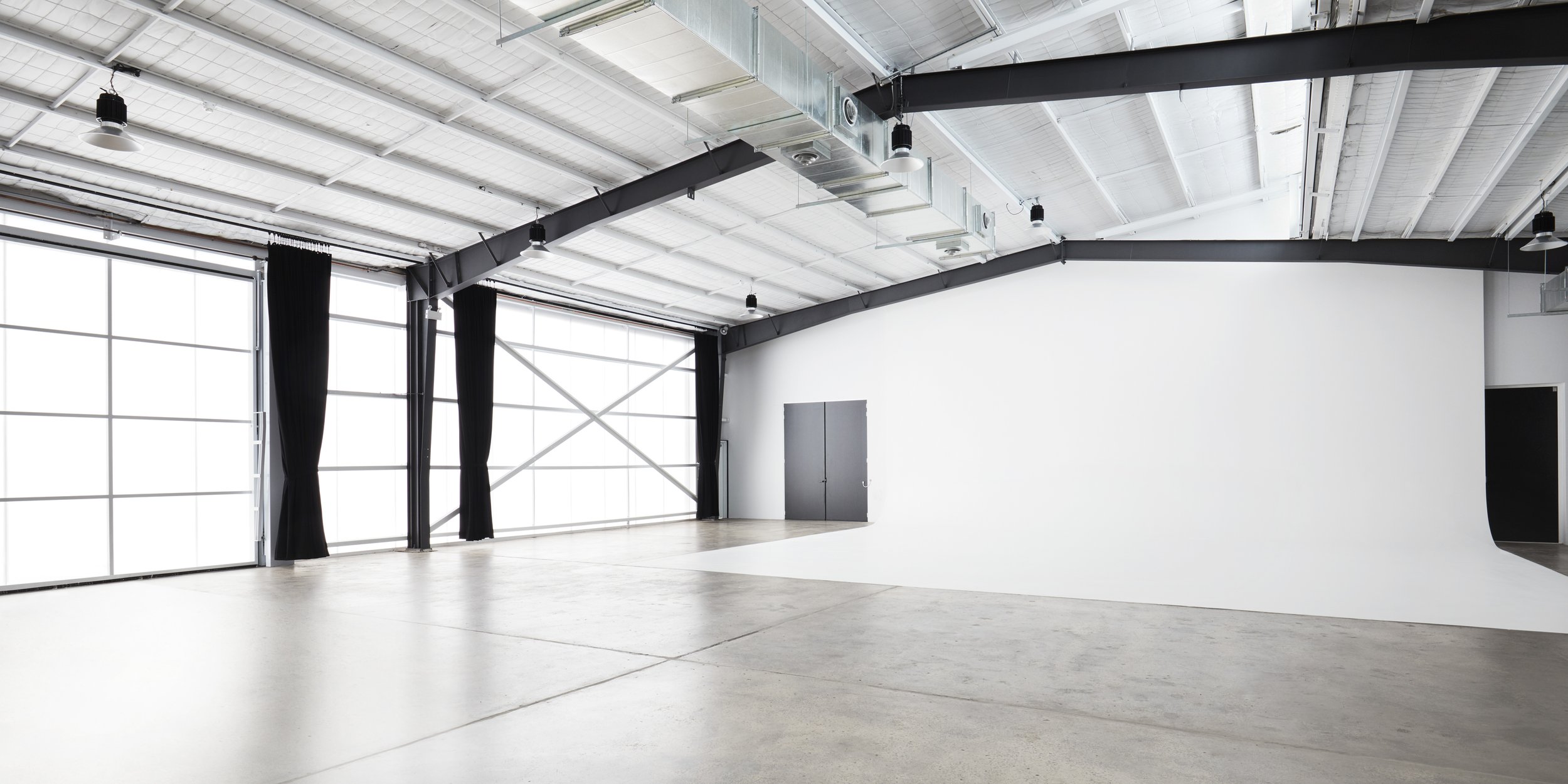 Large, empty photo studio with white cyclorama wall, concrete floor, metal ceiling and frame. Natural and artificial lighting.