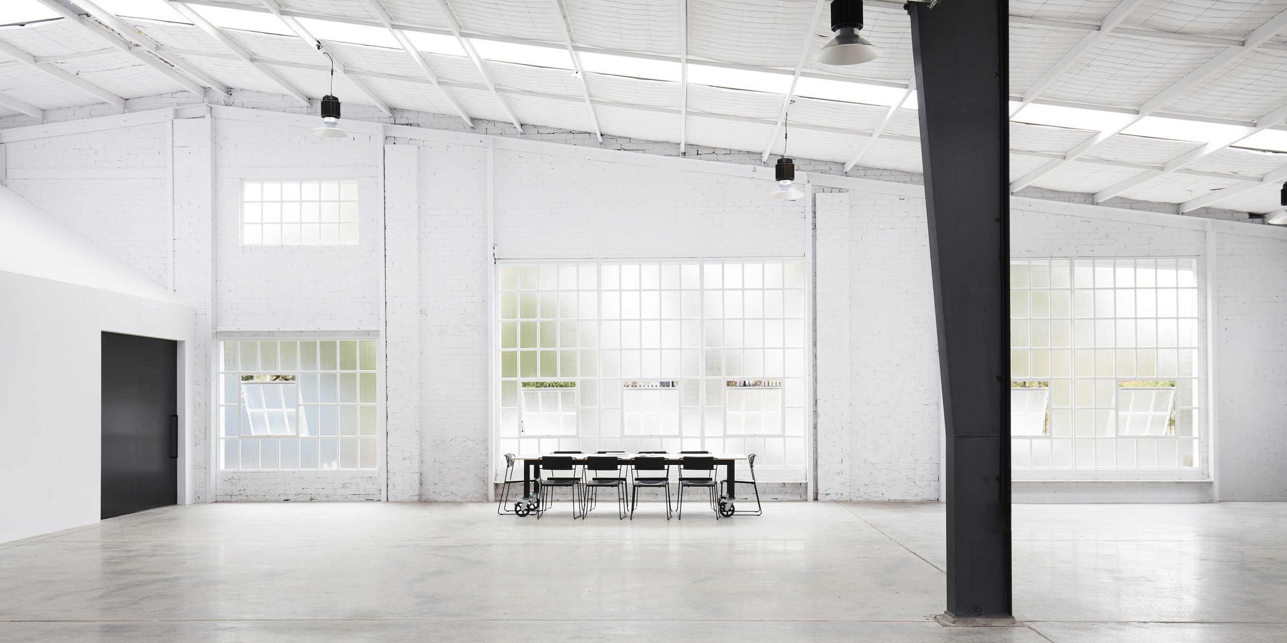Spacious industrial interior with white walls, high ceilings, a long conference table with chairs, and natural light from large windows.