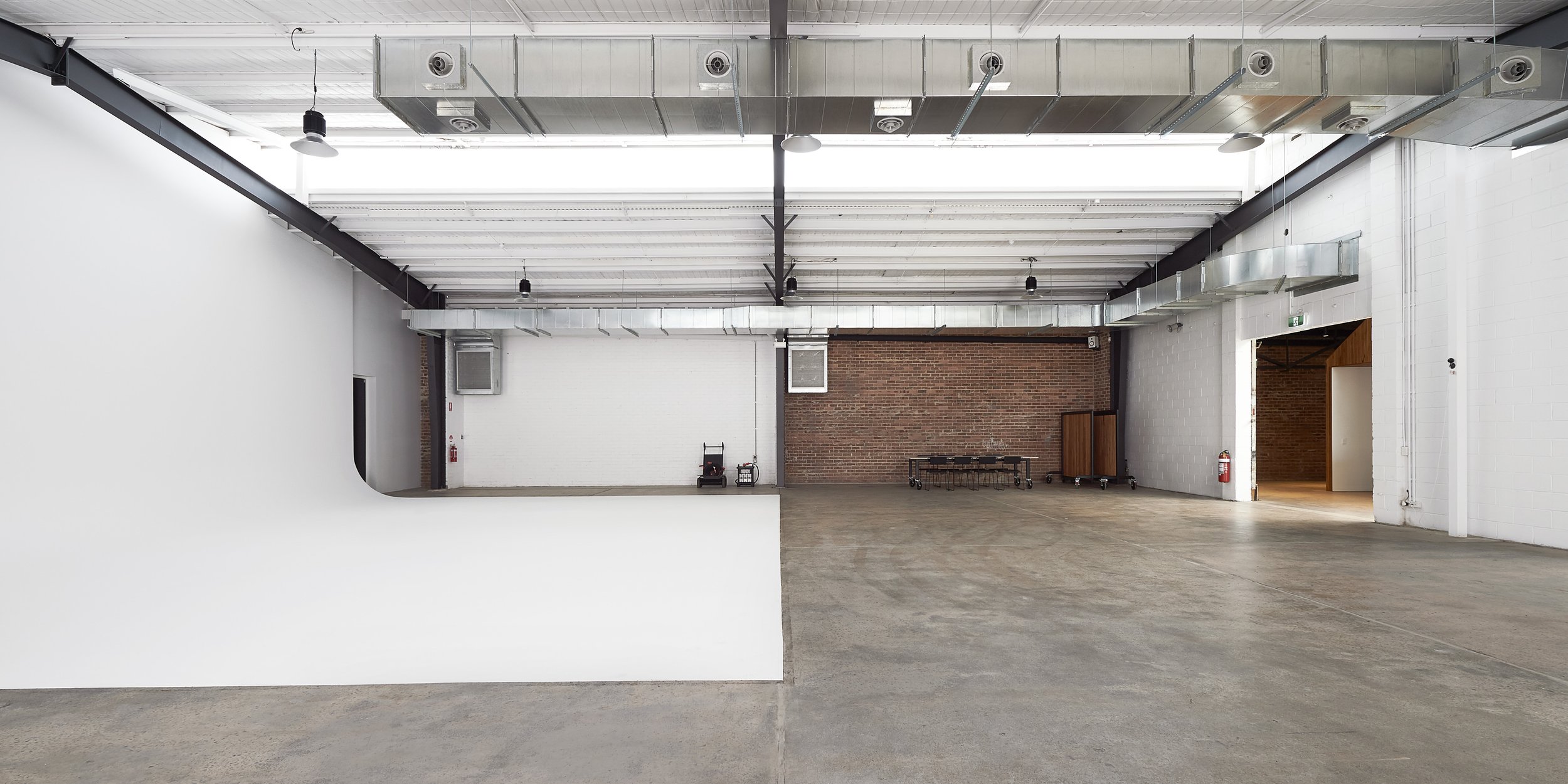 Wide shot of a large, empty photo studio with concrete floors, white walls and a red brick wall; overhead ducting and lighting.