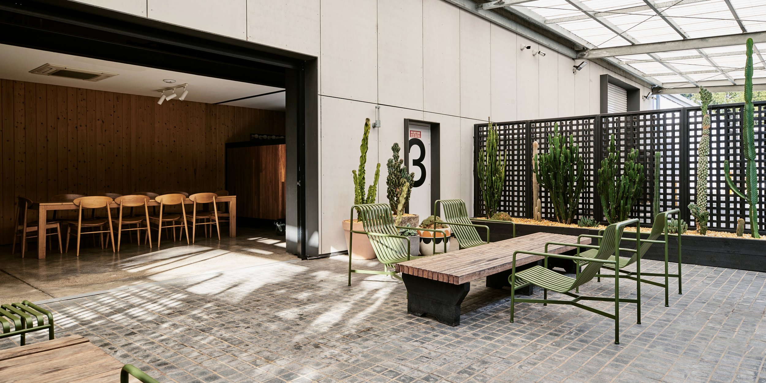 Interior view of a modern atrium with green metal chairs and tables, cacti, and a visible meeting room with a conference table and chairs.