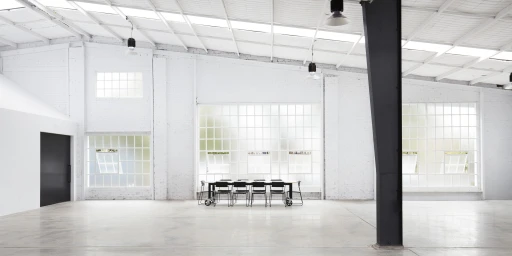 Spacious white photo studio interior with high ceilings, large windows, and a central table with chairs