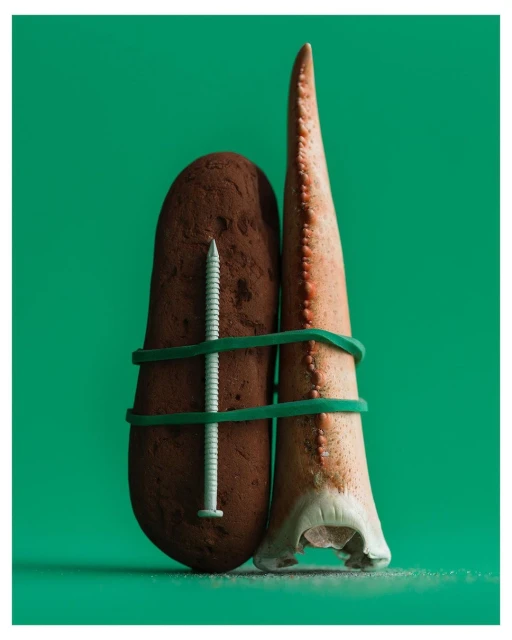 Studio shot of an oddly arranged still life featuring a potato, a screw, and an animal tooth tied together with green rubber bands against green backdrop.