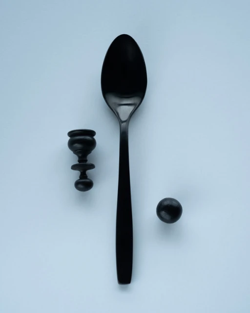 A sleek black spoon lies next to a small ornamental object and dark spherical object on a soft light blue surface, minimalist composition.