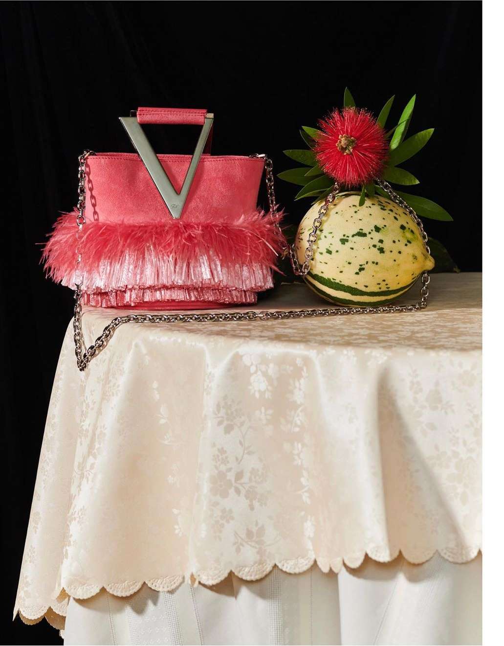 Coral-colored, feathered handbag with chain strap displayed on a table next to a round, green squash and a red flower.