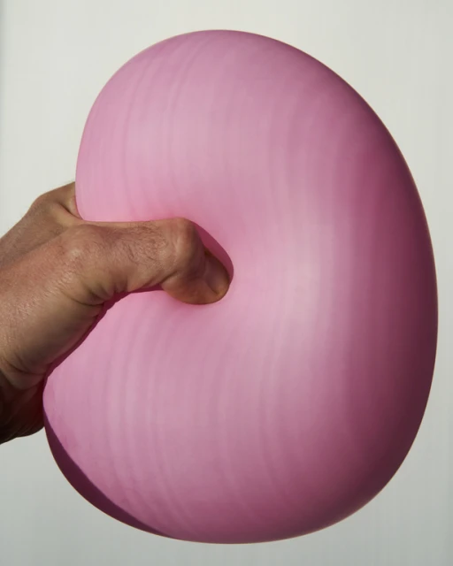 Close-up of a hand squeezing a pink balloon against a white background, showcasing texture and form under studio lighting.