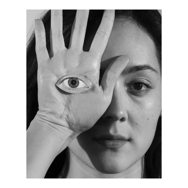 Black and white portrait of a woman with an eye superimposed on her hand covering half of her face in a surreal effect.