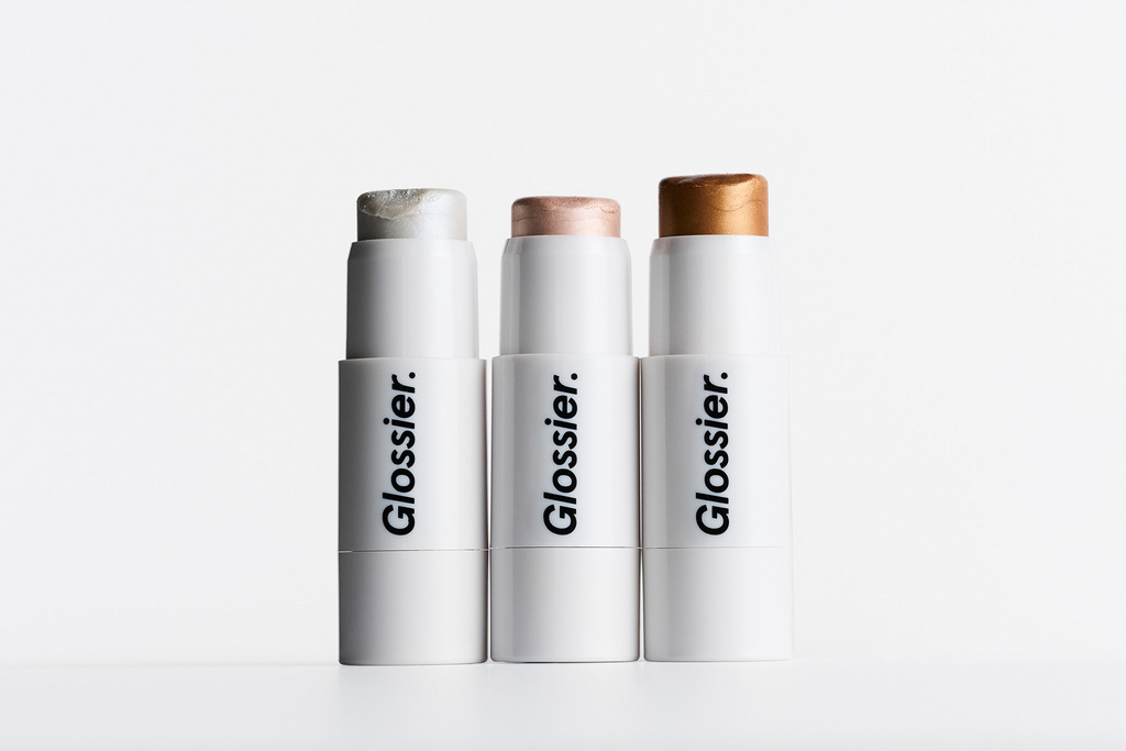 Three Glossier brand highlighting sticks in silver, pink, and gold hues stand side-by-side against a bright white background.