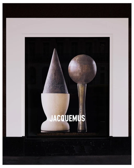 Still life with geometric objects in a window display, featuring a cone and sphere, with the Jacquemus logo in white text.