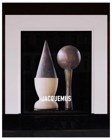 Still life with geometric objects in a window display, featuring a cone and sphere, with the Jacquemus logo in white text.