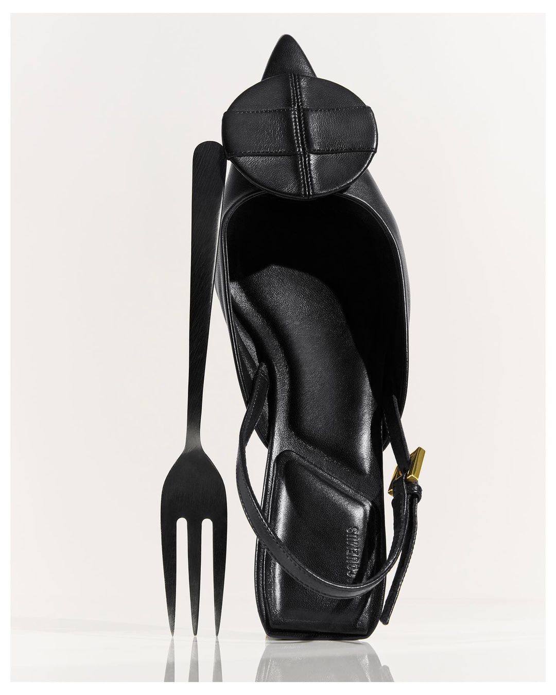 A black leather heeled shoe sits with a black fork against a white backdrop, highlighting the unique juxtaposition of fashion and dining.