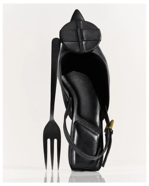 A black leather heeled shoe sits with a black fork against a white backdrop, highlighting the unique juxtaposition of fashion and dining.