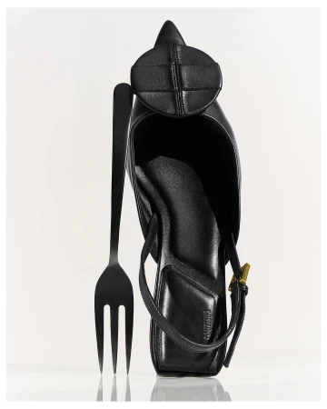 A black leather heeled shoe sits with a black fork against a white backdrop, highlighting the unique juxtaposition of fashion and dining.