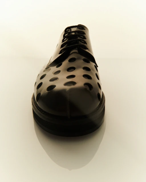 Close-up shot of a black leather-like dress shoe with a round perforated pattern and black laces, placed on a reflective white surface.