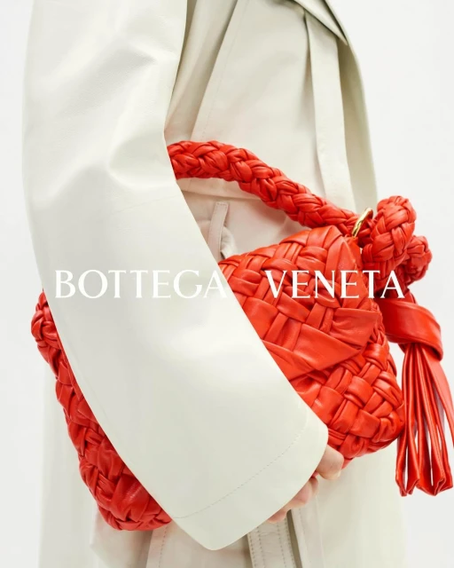 Close-up studio shot of a red braided Bottega Veneta leather handbag carried by a person in an off-white trench coat.