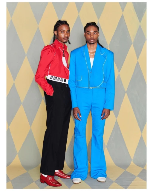 Two adult men in colorful suits pose against a diamond patterned backdrop, one in red with a piano keyboard, the other in bright blue.