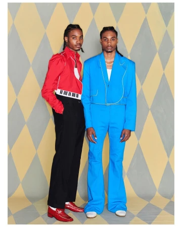 Two adult men in colorful suits pose against a diamond patterned backdrop, one in red with a piano keyboard, the other in bright blue.