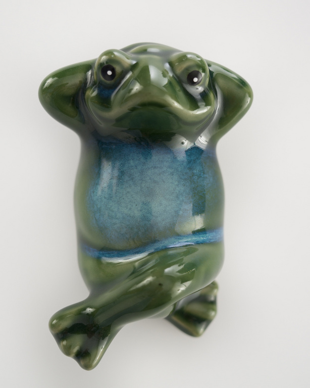 Ceramic frog figurine with hands behind its head, green and teal glaze, perched against a plain, light backdrop in a studio shot.