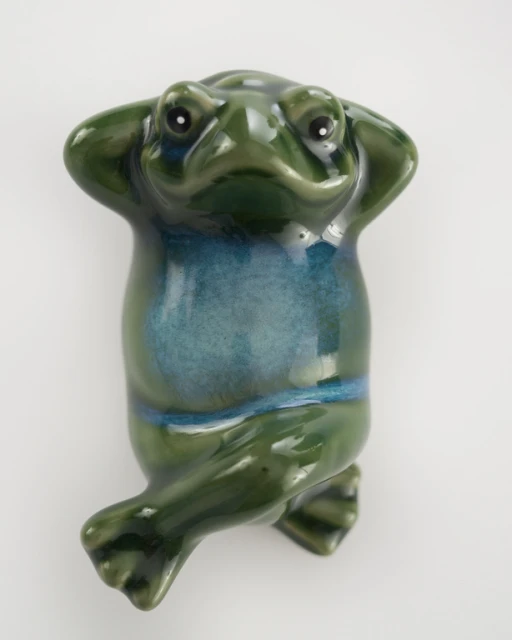 Ceramic frog figurine with hands behind its head, green and teal glaze, perched against a plain, light backdrop in a studio shot.
