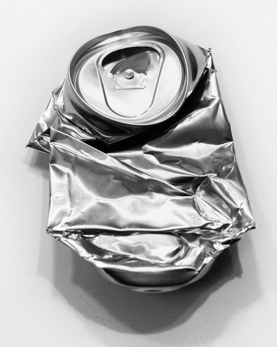 Crushed aluminum can on a white surface. The beverage container is heavily dented with a prominent top with its opening still visible.